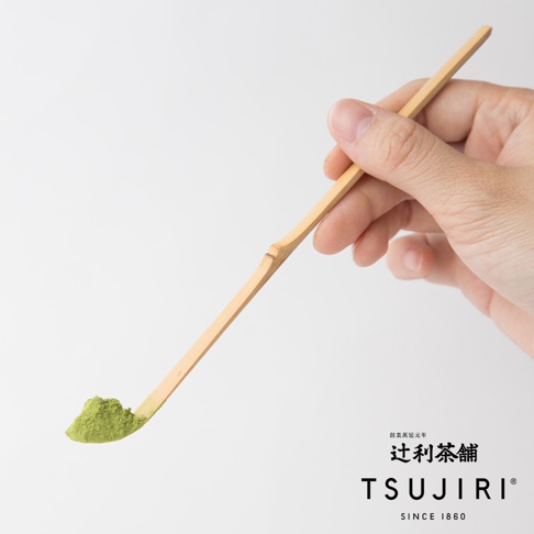 一隻手正持著竹製抹茶勺，勺端盛有一小堆細緻的綠色抹茶粉，畫面右下方印有辻利茶舖品牌名稱與創始年份標誌，背景為簡潔明亮的純白色。