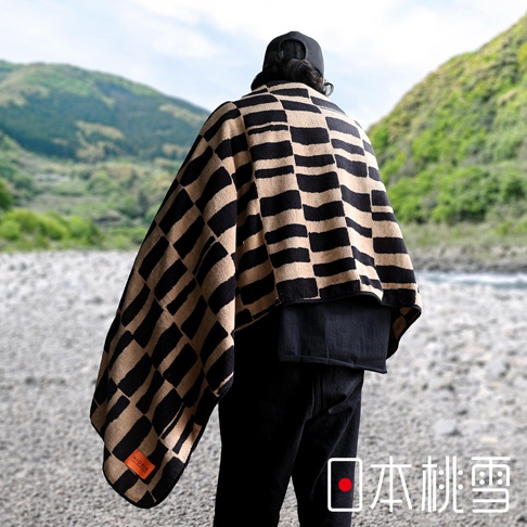 日本桃雪 logo 標誌的戶外格紋棉柔浴巾，一人背部視角，置於河邊的礫石場地上