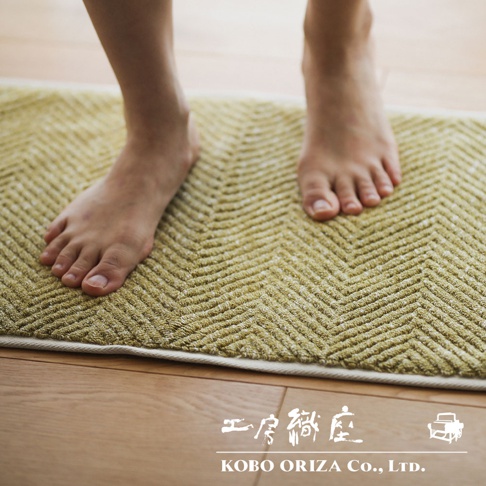 一雙赤腳踩在淡黃色棉麻混紡人字紋浴墊上，浴墊邊緣為白色滾邊，背景為木質地板。