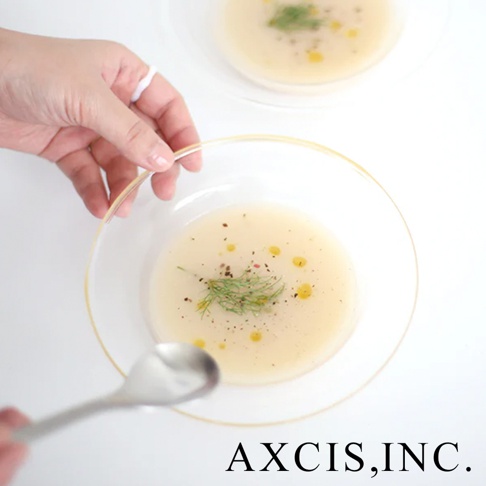 一個裝有奶油湯、點綴著蒔蘿和黑胡椒的透明玻璃碗，碗緣有金色裝飾。一隻手正用湯匙舀湯。