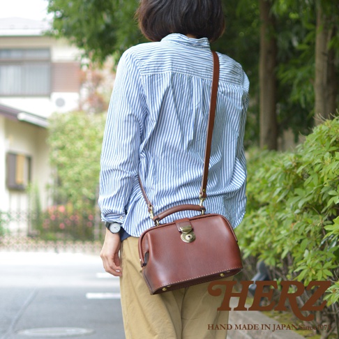職人手作皮革方型迷你醫生包，深棕色皮革，配有金色金屬扣和可調式肩帶，由一位身穿藍白條紋襯衫的人士攜帶，在戶外拍攝。