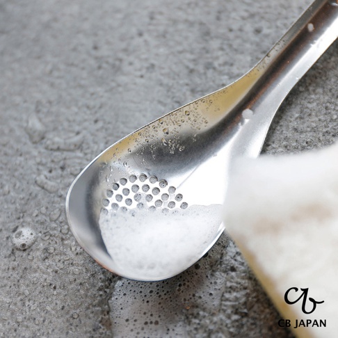CB JAPAN 日本製不鏽鋼瀝汁匙，水珠沾附在金屬勺面上，勺內有泡沫，背景是灰色紋理桌面。
