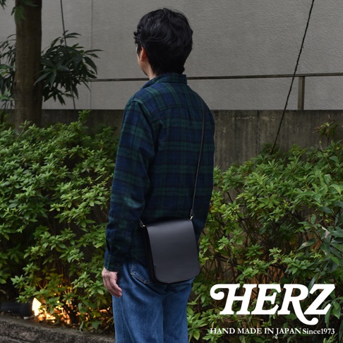 一位男性背著日本HERZ職人手作黑色皮革直式單肩包，側面展示其簡約的翻蓋設計與皮革質感，背景為綠色植栽，畫面右下角印有品牌標誌與手工製造字樣。
