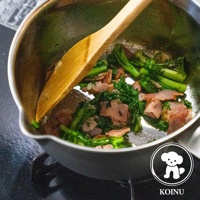 三層不鏽鋼雪平鍋內，木鏟正在翻炒綠色蔬菜和切塊的培根。鍋身為銀色不鏽鋼材質，鍋柄為灰色。