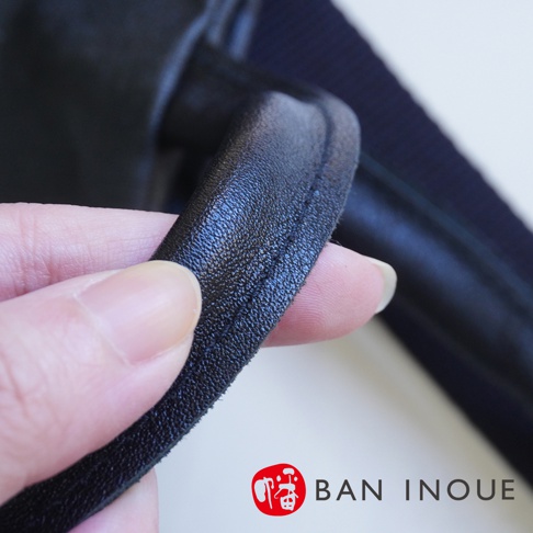 BAN INOUE 標誌旁的一塊黑色皮革，手指正在觸摸皮革的縫線細節