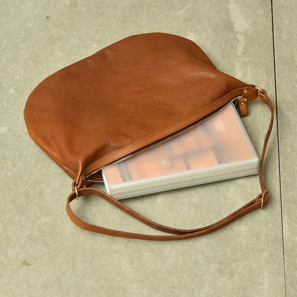 職人手作皮革Organ單肩半月包，採用棕色柔軟皮革製成，包身半月形，帶有拉鍊開口，露出內部裝有淺色文件收納夾。