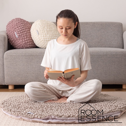 一位女士坐在圓形涼感夾心包覆圓墊上閱讀。圓墊為大地色系，表面有立體紋理。背景為灰色沙發，上方擺放著粉色和米白色的圓形靠墊。