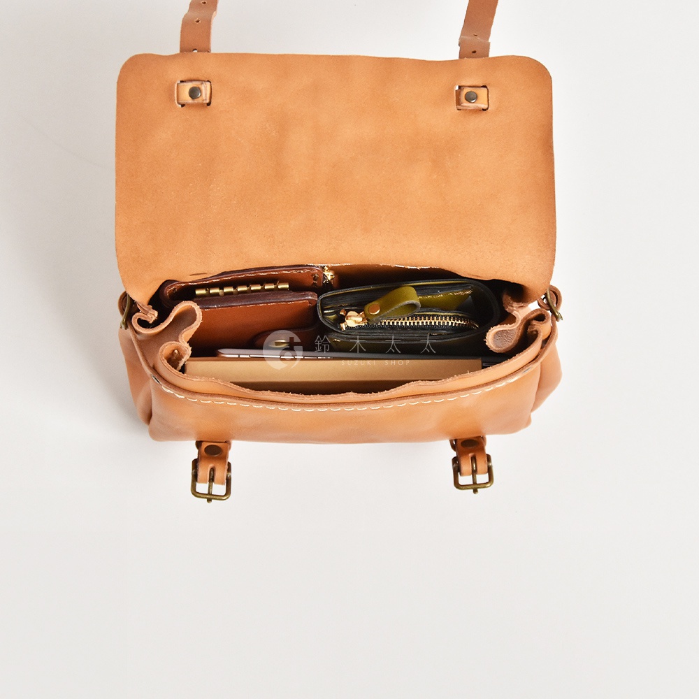 復古棕色皮革郵差包，打開狀態，內部裝有錢包、卡夾和筆記本。包身有手工縫線細節及金屬扣環。