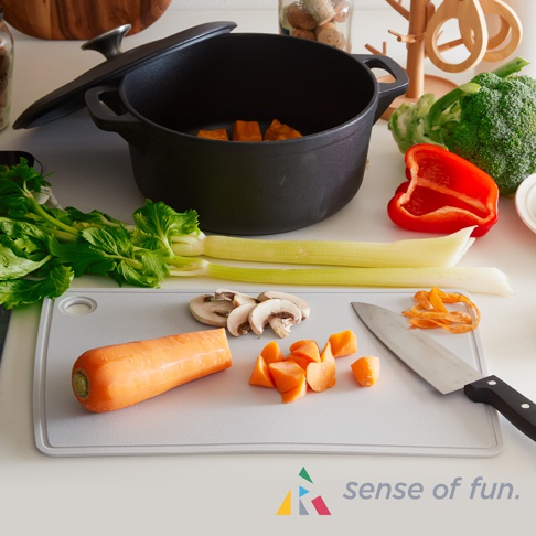 淺灰色矽膠砧板上，擺放著一根完整的胡蘿蔔、切片蘑菇、切塊胡蘿蔔、芹菜、半顆紅甜椒、一株西蘭花，以及一個黑色鑄鐵鍋，鍋內有切塊南瓜。