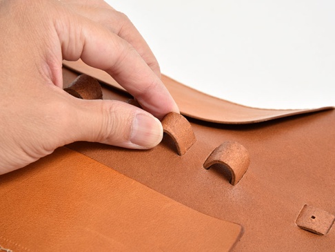 職人手作皮革捲捲收納筆袋，展示棕色牛皮材質，特寫手指正在操作皮革束帶，展現產品細節與質感。