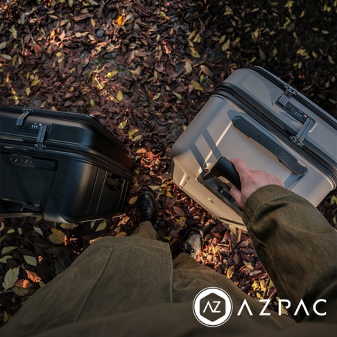 第一人稱視角拍攝，一個人穿著橄欖綠色大衣手持淺灰色 AZPAC 20 吋前開口煞車行李箱，左側地面同時放置一個黑色行李箱，背景為鋪滿秋季枯黃落葉的地面。