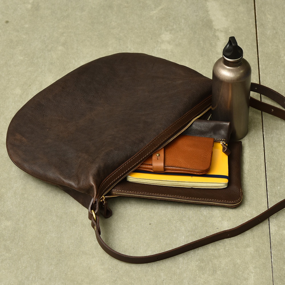 深棕色皮革半月型單肩包，包內裝有筆記本、棕色皮夾和銀色水瓶。
