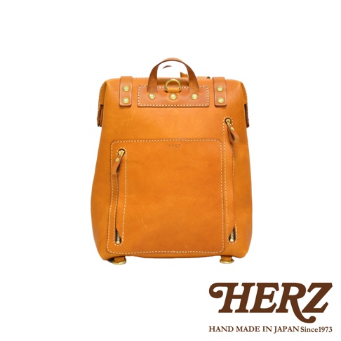 職人手作皮革後背包 R-143，採用溫暖的棕褐色皮革，設計簡約，正面有兩個拉鍊口袋，頂部有提把和金屬扣環，展現精緻工藝。