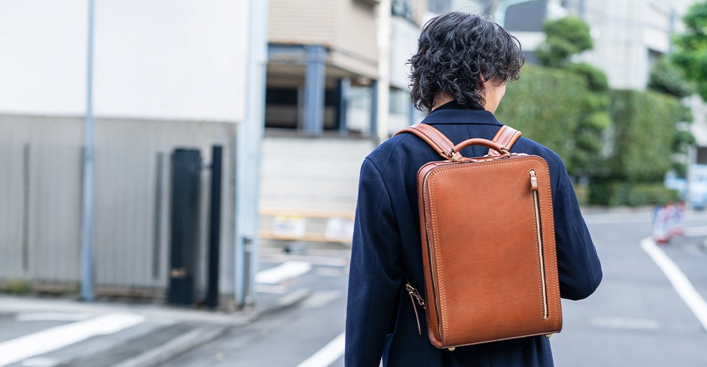 棕色皮革製的職人手作商務電腦後背包，背包側面有拉鍊開口，肩帶與提把均為皮革材質。