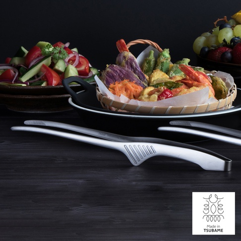 一對銀色金屬材質的公筷夾，筷夾前端有防滑紋路，另一雙為黑色。背景有炸物籃和沙拉。