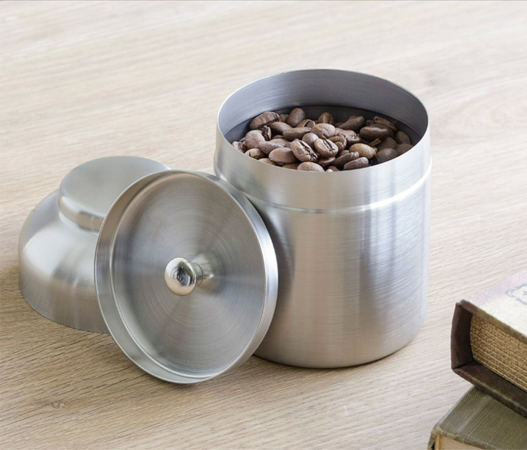 日製18-8不鏽鋼儲豆罐，附帶一個銀色不鏽鋼製的量匙，罐身裝有淺棕色咖啡豆，蓋子朝上放置於罐旁，整體呈現銀色金屬光澤。