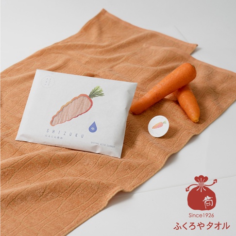 一條橘色毛巾，表面有壓紋，上面放著一個印有胡蘿蔔圖案的白色包裝袋，袋子下方標示「SHIZUKU DYING WITH CARROT」，旁邊有兩根新鮮胡蘿蔔和一個圓形貼紙。