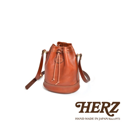 職人手作皮革束口單肩包，呈現自然棕色，以紮實縫線工藝打造，束口處有細緻的綁繩設計，質感厚實的皮革散發復古韻味，配有可調整長度的肩背帶。