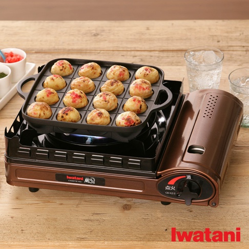 銅色金屬機身卡式爐，上方放置著一個黑色方形烤盤，烤盤內有 16 個金黃色的章魚燒，部分章魚燒表面點綴著紅色醬料。