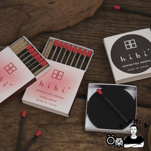 台灣限定版香氛火柴組合，包含三個紙盒裝的火柴，火柴頭為紅色，火柴棒為黑色。其中兩個紙盒為粉紅色漸層，印有白色方格圖案及「hibi」品牌字樣。另一個圓形黑底白字包裝盒，以及一個白色小盒，內有一個黑色圓形墊片和一根火柴。