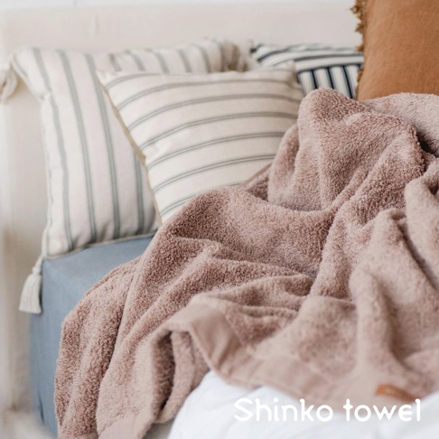 MAISON BLANC 輕奢柔膚匹馬棉浴巾，顏色為柔和的粉棕色，質地蓬鬆柔軟，呈現細膩的毛圈紋理。浴巾旁擺放著數個條紋抱枕，營造舒適居家氛圍。