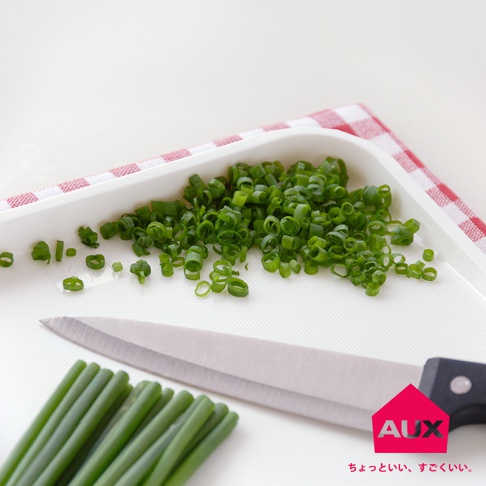 白色防溢大砧板上，部分蔥段被切成細圈，旁邊擺放著未切的青蔥段和一把不鏽鋼刀。背景為粉紅色格紋布。