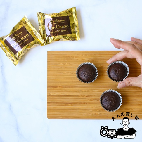 金黃色包裝的烤巧克力菓子禮盒，內含三個獨立包裝的巧克力菓子，以及放在木質砧板上的三個巧克力菓子，其中一個正被手拿起。