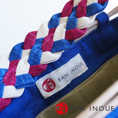 BAN INOUE 麻花辮束口手提包，展示了其獨特的編織工藝，藍色、白色和桃紅色的織帶交織成精緻的麻花辮，搭配藍色帆布包身，標籤上印有品牌名稱 BAN INOUE 及日文標誌。