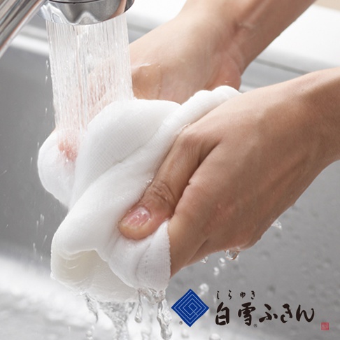 一雙手正在水槽中沖洗一塊白色的木纖維拭巾。拭巾材質柔軟，水流從水龍頭流下，浸濕拭巾並滴落。