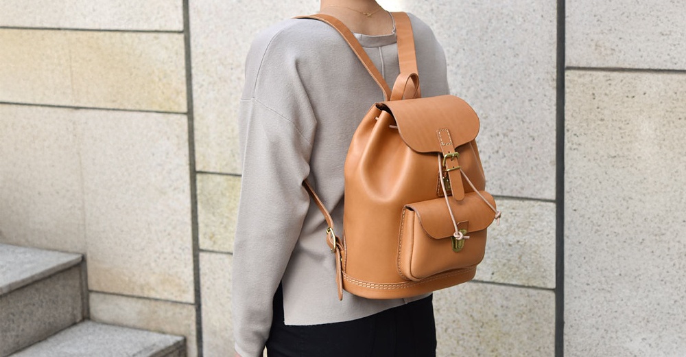 淺棕色皮革後背包，具有抽繩閉合、翻蓋和前置小口袋，搭配金屬扣環及綁帶，背包側面有縫線裝飾。