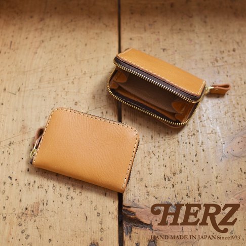 兩款職人手作皮革收納錢包，皆為淺棕色，材質為牛皮。其中一款錢包已拉開拉鍊，展現內部多層隔間設計。另一款錢包闔上，表面光滑，邊緣縫線明顯，並有金色拉鍊及皮革拉環。