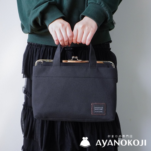 一位身穿綠色毛衣和黑色荷葉邊裙子的女士，雙手提著一個黑色橫式口金手提包，包上有一個方形的品牌標籤，下方是「AYANOKOJI」的品牌名稱和標誌。