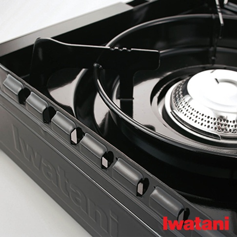 黑色金屬材質的卡式爐，配有黑色爐架和銀色爐頭，爐身側面有「Iwatani」字樣和防風結構的開孔設計。