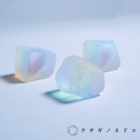三顆半透明的合光稜鏡石，呈現柔和的彩虹光澤，有立方體和不規則多面體的形狀，材質看似玻璃或水晶，散發著淡藍、粉紅和淡黃色的光芒。