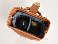 職人手作皮革兩用迷你口金單肩包 (CW-7)，以棕色皮革製成，包內有灰色襯裡，並可容納一個 Nikon 相機鏡頭、一個綠色吹塵球和一個黑色收納袋。