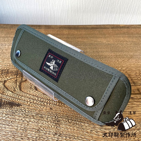 深綠色純棉帆布製成的長形收納包，正面有黑色方形標籤，印有白色犬隻圖案及日文文字。收納包邊緣有細緻的白色縫線，並以銀色金屬釦固定。