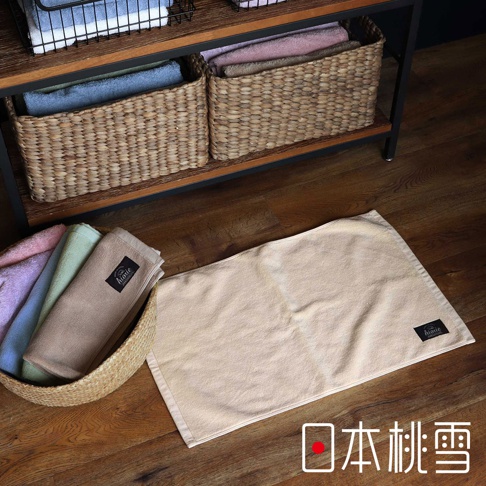 一張淺米色長方形浴墊，材質為柔軟的毛巾布，角落有黑色品牌標籤。一旁竹編籃中疊放著其他顏色的浴巾，包含淺紫、藍、綠及卡其色。