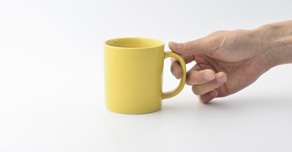 一只手拿著一個黃色的陶瓷馬克杯，杯身簡潔圓潤，帶有一個弧形把手。