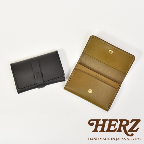 日本職人品牌HERZ手作皮革名片夾，左側為黑色款呈現閉合狀態，右側為橄欖綠色款呈現打開狀態，展示出內部夾層與金屬按扣設計，整體風格簡約復古，下方印有HERZ品牌商標與創立年份資訊。