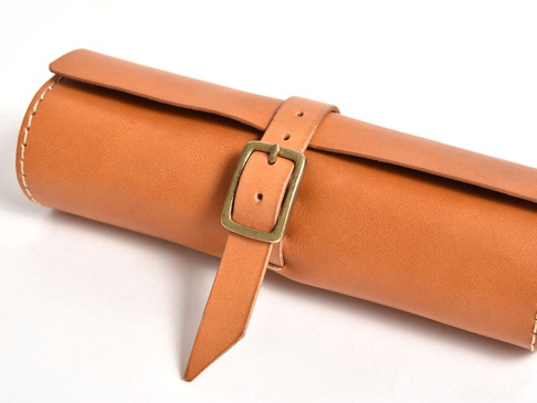 職人手作皮革捲捲收納筆袋，採用天然棕色皮革，搭配仿舊銅色金屬釦環，側邊以米白色線跡縫製，設計簡約實用。