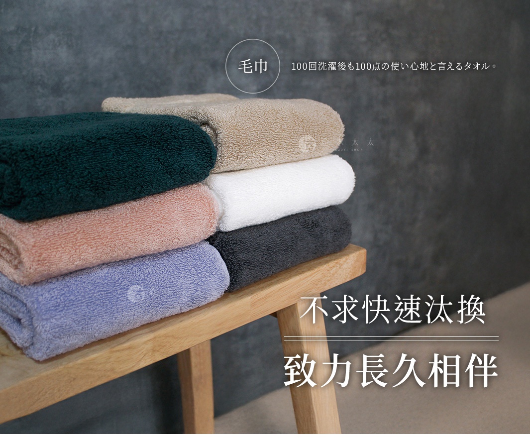 一疊今治毛巾，顏色包含深綠、粉紅、淺紫、米白、白色和炭灰色，質地柔軟，陳列在木製層架上。