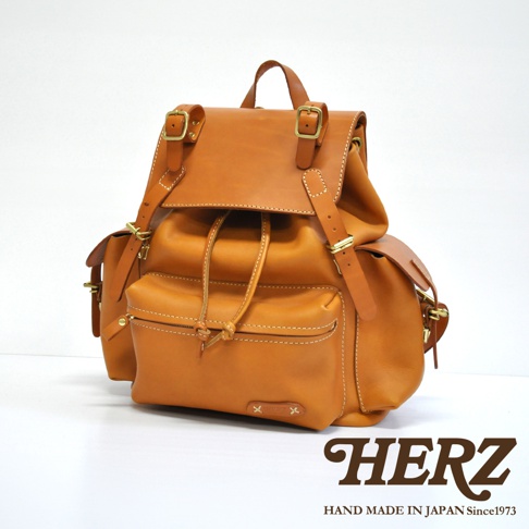 職人手作皮革復古風旅行後背包，採用溫暖的棕褐色皮革，配有黃銅色金屬扣環和抽繩閉合。包款設有翻蓋主袋、抽繩束口以及前置拉鍊口袋和側邊口袋，整體風格簡約實用。