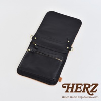 職人手作皮革翻轉雙色直式斜背包，主體為黑色皮革，翻蓋內側及斜背包外層下方為原色皮革，設有拉鍊小袋及金屬掛環。