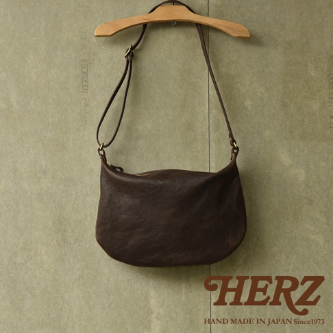 職人手作皮革Organ單肩半月包，深棕色，材質為柔軟皮革，包身呈彎月形，配有可調節長度的細長肩帶，以拉鍊開合，掛於木質衣架上，背景為淺綠色紋理牆面。