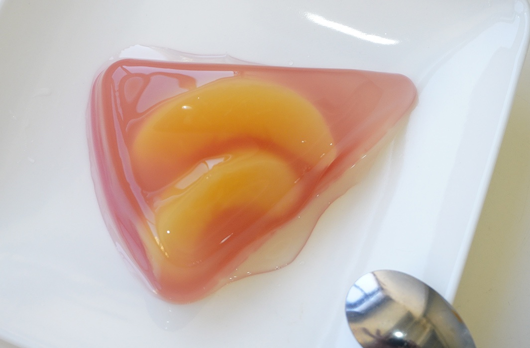 三角形果凍內含兩片桃子，果凍呈現粉紅色，質地光滑，盛裝於白色盤中，盤旁有一支湯匙。