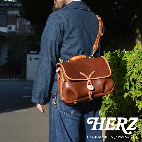 棕色皮革製郵差包，採用鹿角扣繩設計，搭配可調式皮革肩帶和黃銅金屬扣環。包包側面有品牌標誌。