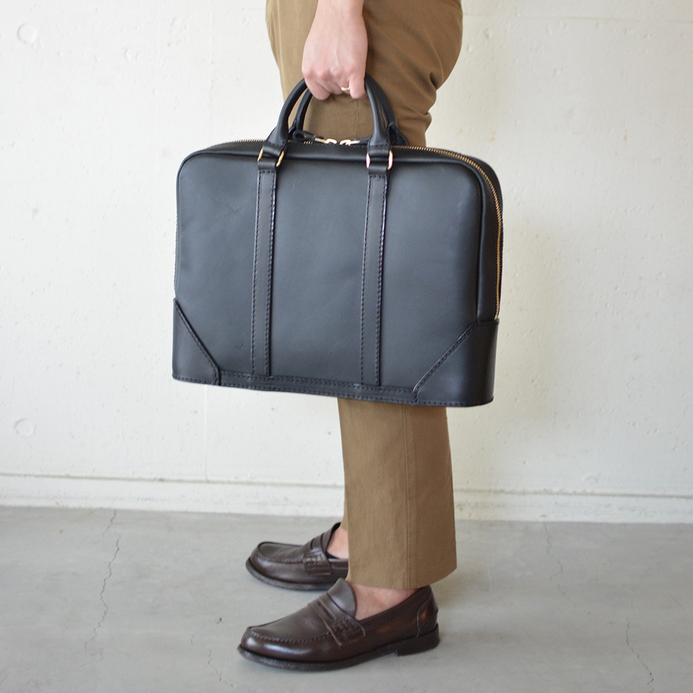黑色皮革商務公事包，配有金色拉鍊和雙提把，由一個人拿著，腳穿著咖啡色樂福鞋。