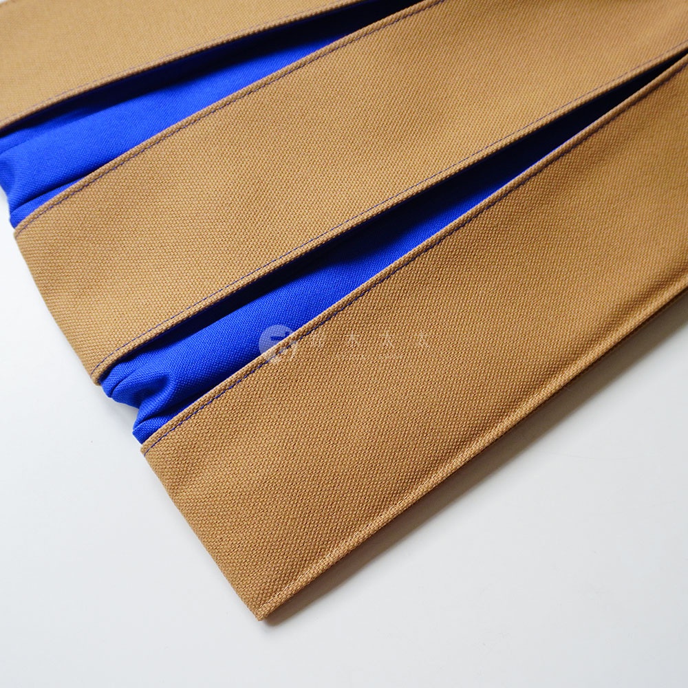 這是一張近距離特寫照片，呈現京都KOSHO品牌的ougi帆布信玄袋細節。畫面由多層厚實的駝色帆布交疊而成，邊緣縫線細緻平整。帆布褶皺間露出內襯鮮豔的藍色織物，強烈的色彩對比突顯出袋身的層次感與堅韌的織物紋理，拍攝角度從上方俯視，強調材質的立體感與工藝品質。