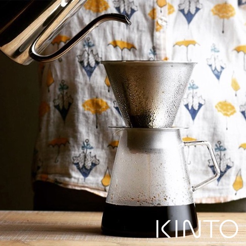 銀色金屬濾網架置於透明玻璃咖啡壺上，壺內盛有深棕色咖啡，背景為印有黃色雨傘圖案的布料。