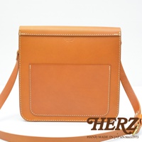 職人手作經典厚實單肩包，採用天然鞣製皮革製成，呈現溫潤的淺棕色。包款設計簡潔，正面有一個方形貼袋，邊緣以白色縫線勾勒，整體造型方正硬挺，附有可調節的長肩帶。